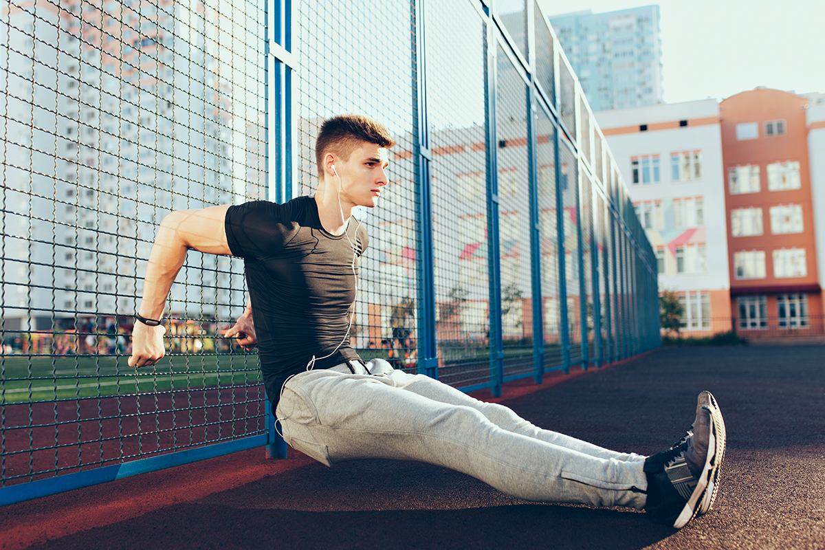 Tense guy with a muscular body at workout training near fence in the morning on stadium. He wears sport clothes, listening to music with headphones. He looks concentrated